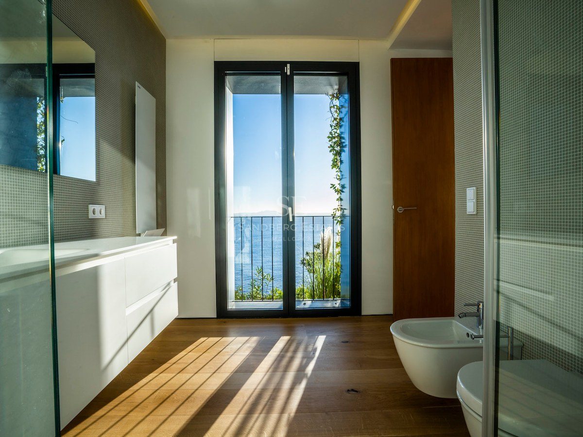 Modern bathroom with oak floors, white vanity, and glass door opening to a balcony with sea views.