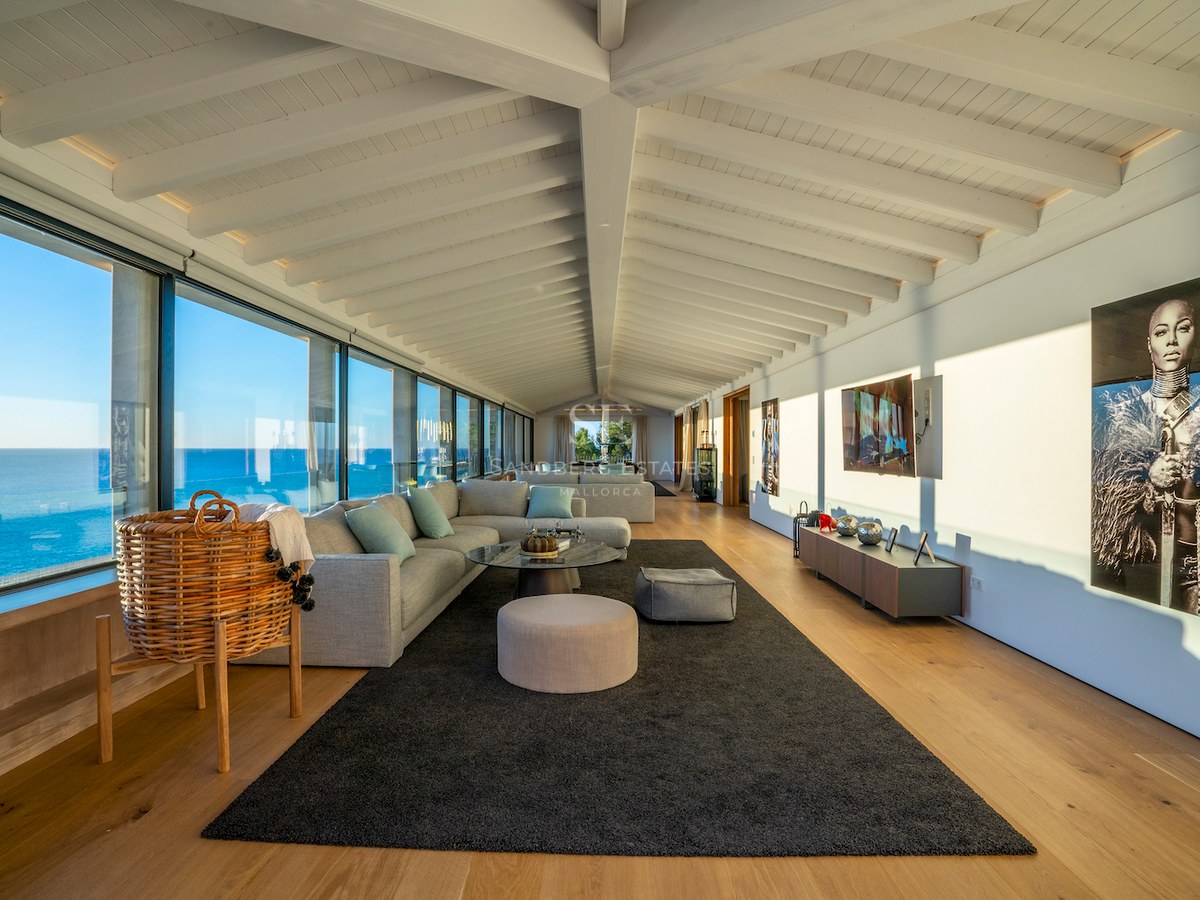 Contemporary living room with white beamed ceilings, oak floors, and floor-to-ceiling windows overlooking the sea.