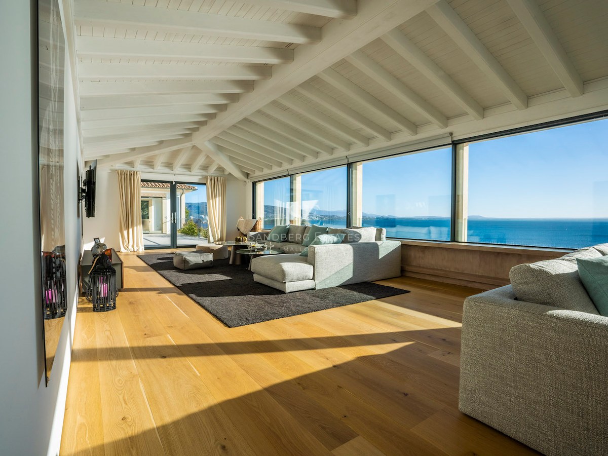 Bright living room featuring large panoramic windows overlooking the sea, white beamed ceilings, and oak wood floors.