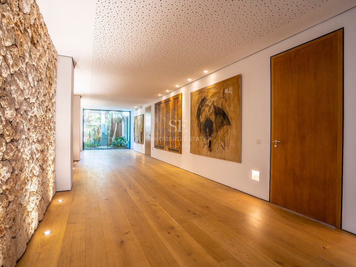 A long modern hallway with wide oak floorboards, white walls with art, and a glass door leading to a garden.