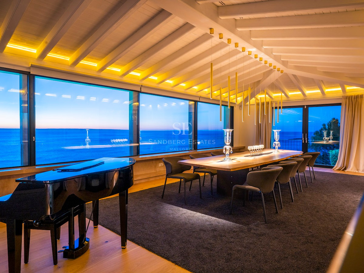 A modern dining room featuring a grand piano, large wooden table, and floor-to-ceiling windows overlooking the sea at dusk.