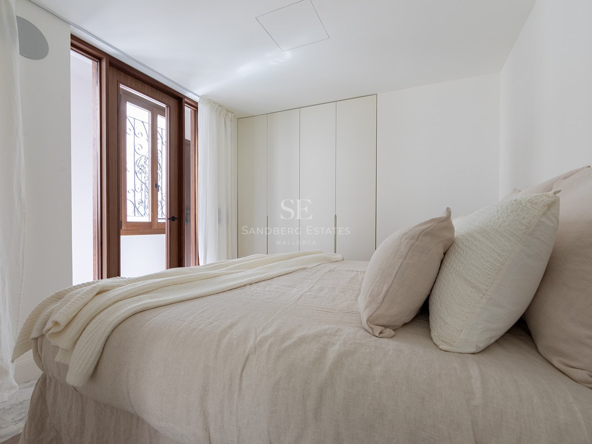 Dormitorio luminoso con ropa de cama de lino beige, armarios blancos minimalistas y puerta de madera al balcón.