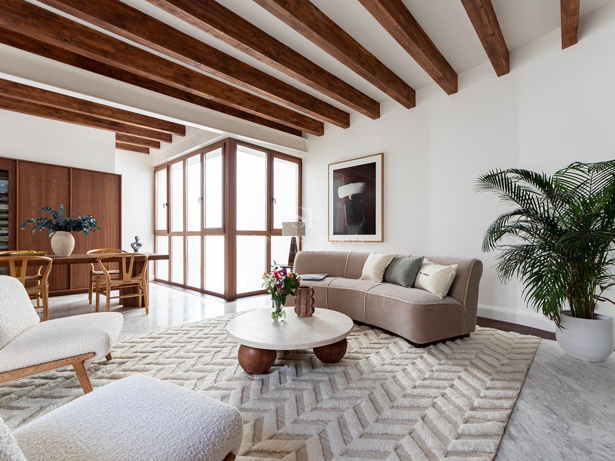 Sala de estar moderna con vigas de madera oscura, suelo de mármol, sofá beige curvo y sillones de borreguillo blanco.