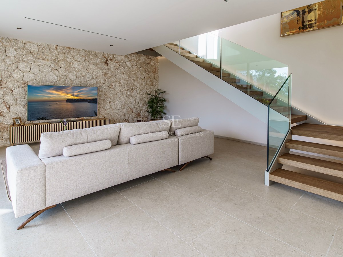 Salon spacieux avec canapé beige, mur en pierre décoratif, télévision et escalier en bois avec garde-corps en verre.