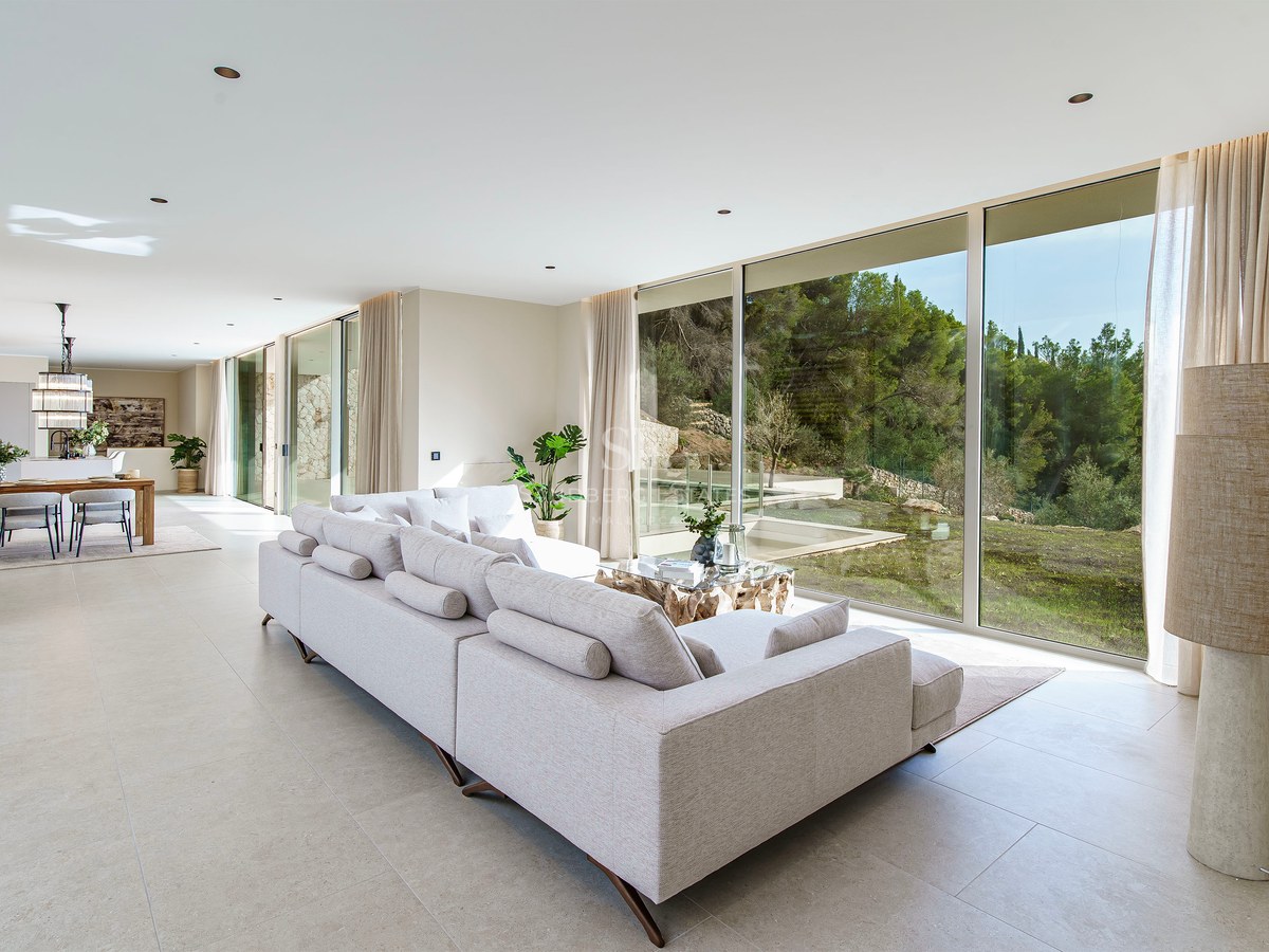 Grand salon moderne avec un canapé gris et des baies vitrées donnant sur une forêt verdoyante.