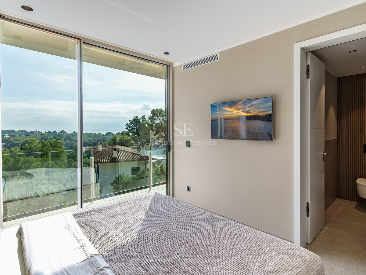 Une chambre moderne dotée d'une grande baie vitrée donnant sur une forêt et la mer, avec une salle de bain attenante.