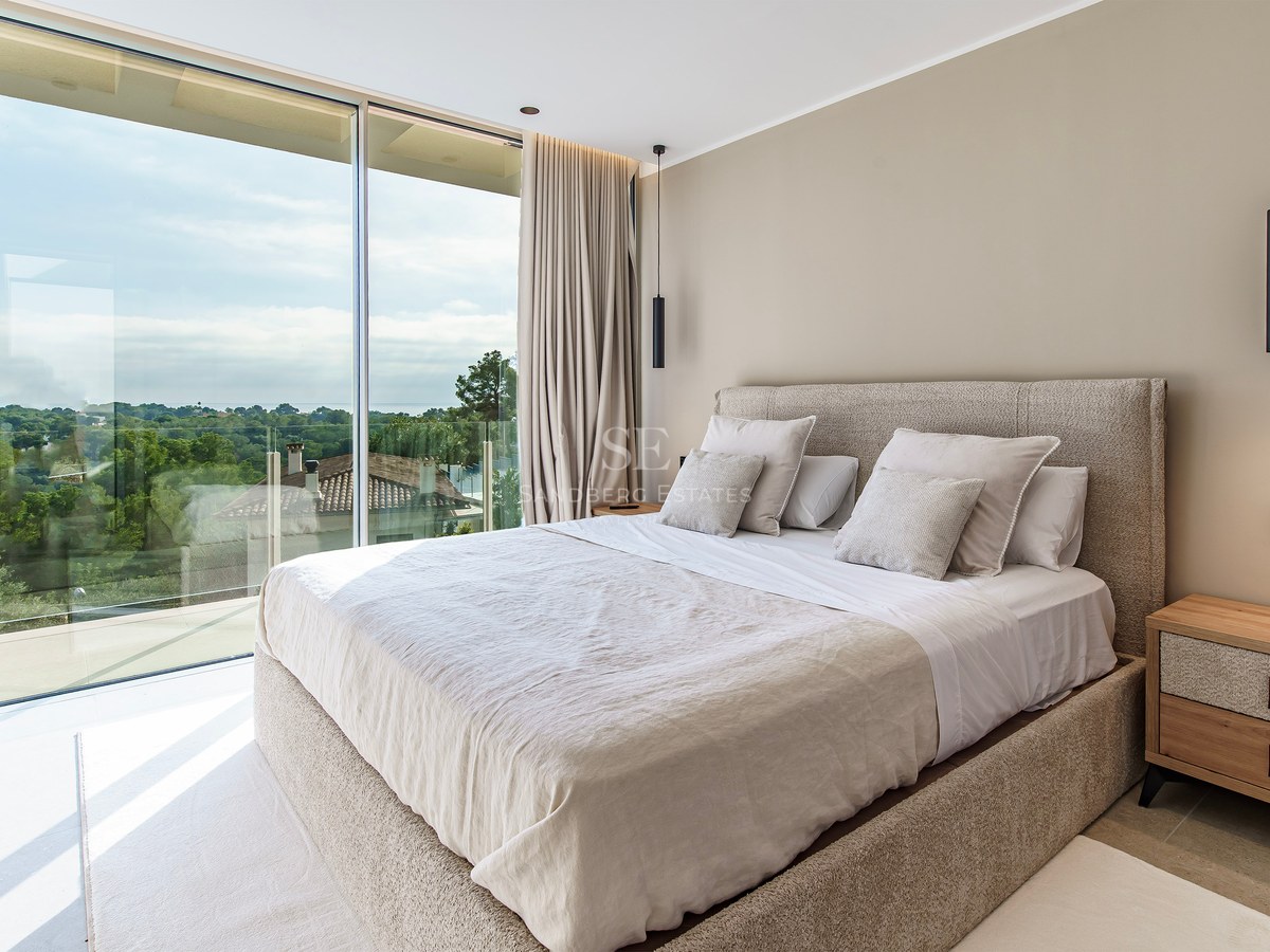 Chambre parentale moderne avec baies vitrées du sol au plafond donnant sur un paysage verdoyant et grand lit neutre.