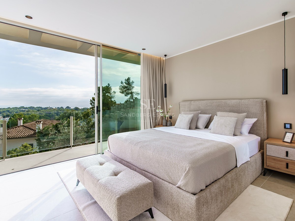 Chambre de luxe avec lit beige et grandes baies vitrées ouvrant sur un balcon avec vue sur la verdure.