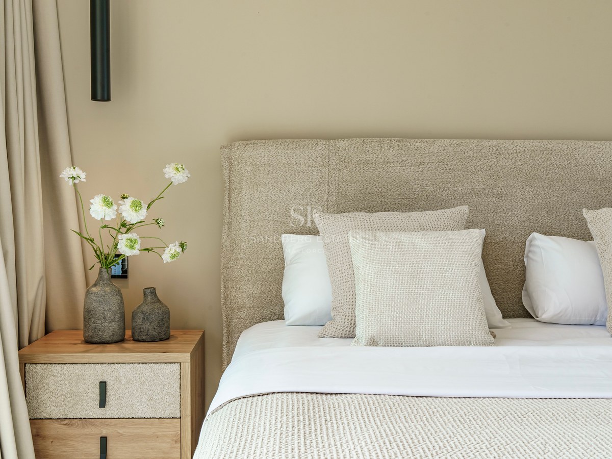 Lit de luxe avec tête de lit texturée, table de chevet en bois et vases décoratifs avec fleurs blanches.