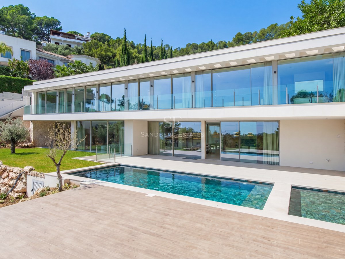 Villa moderne blanche de deux étages avec de grandes baies vitrées face à une piscine rectangulaire et terrasse en bois.