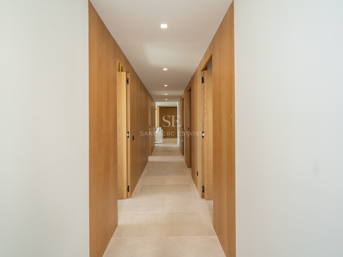 Long couloir moderne avec murs en bois clair, portes invisibles intégrées et sol en pierre beige sous spots LED.