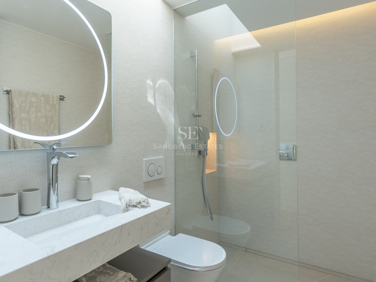 Modern bathroom featuring a circular LED mirror, marble vanity, and glass walk-in shower under a skylight.