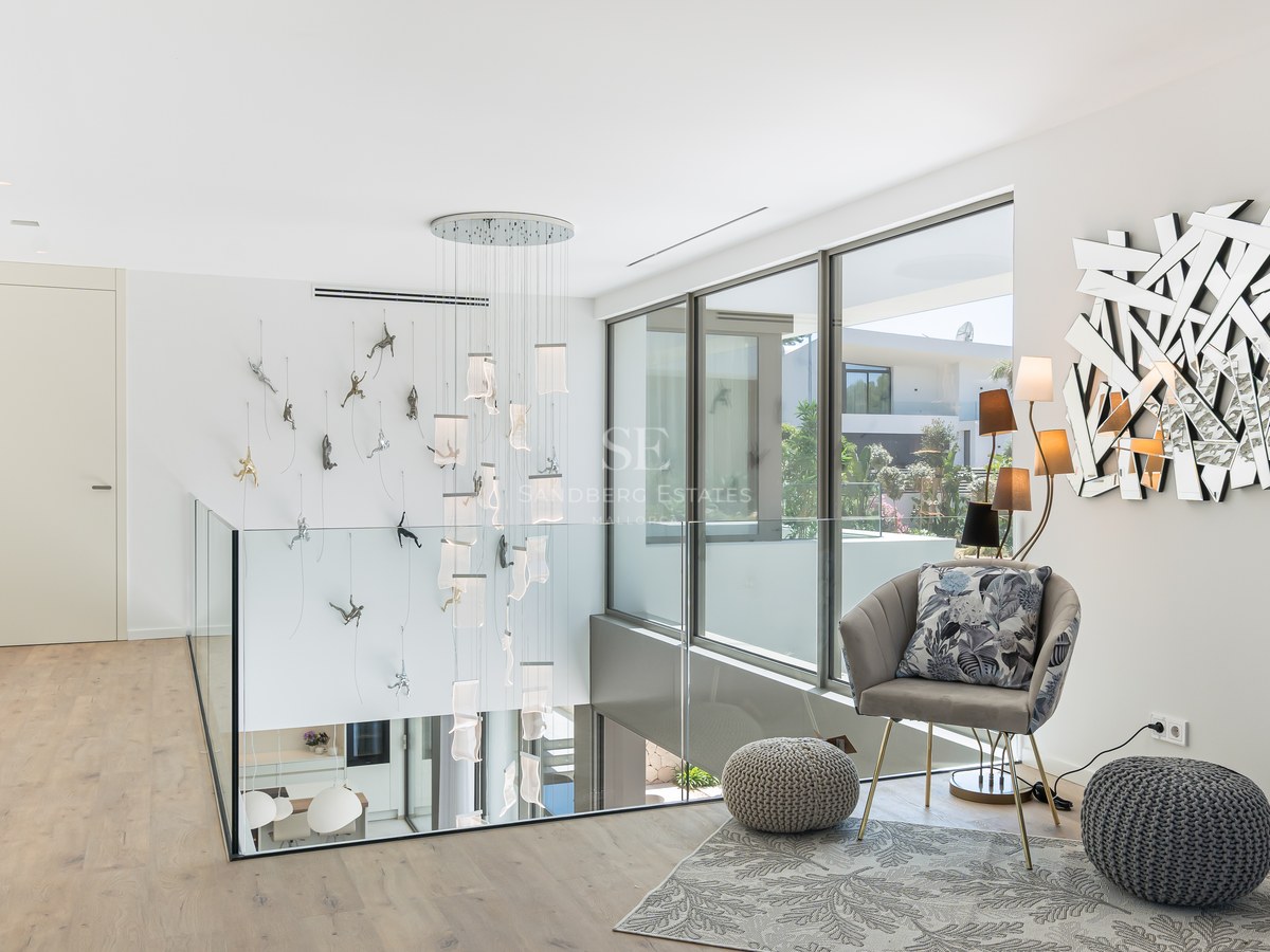 A contemporary hallway landing with glass railings, a tall cascading chandelier, and floor-to-ceiling windows.
