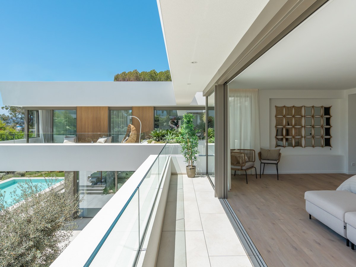 High-angle view of a modern master bedroom opening onto a white stone terrace with glass railings overlooking a pool.