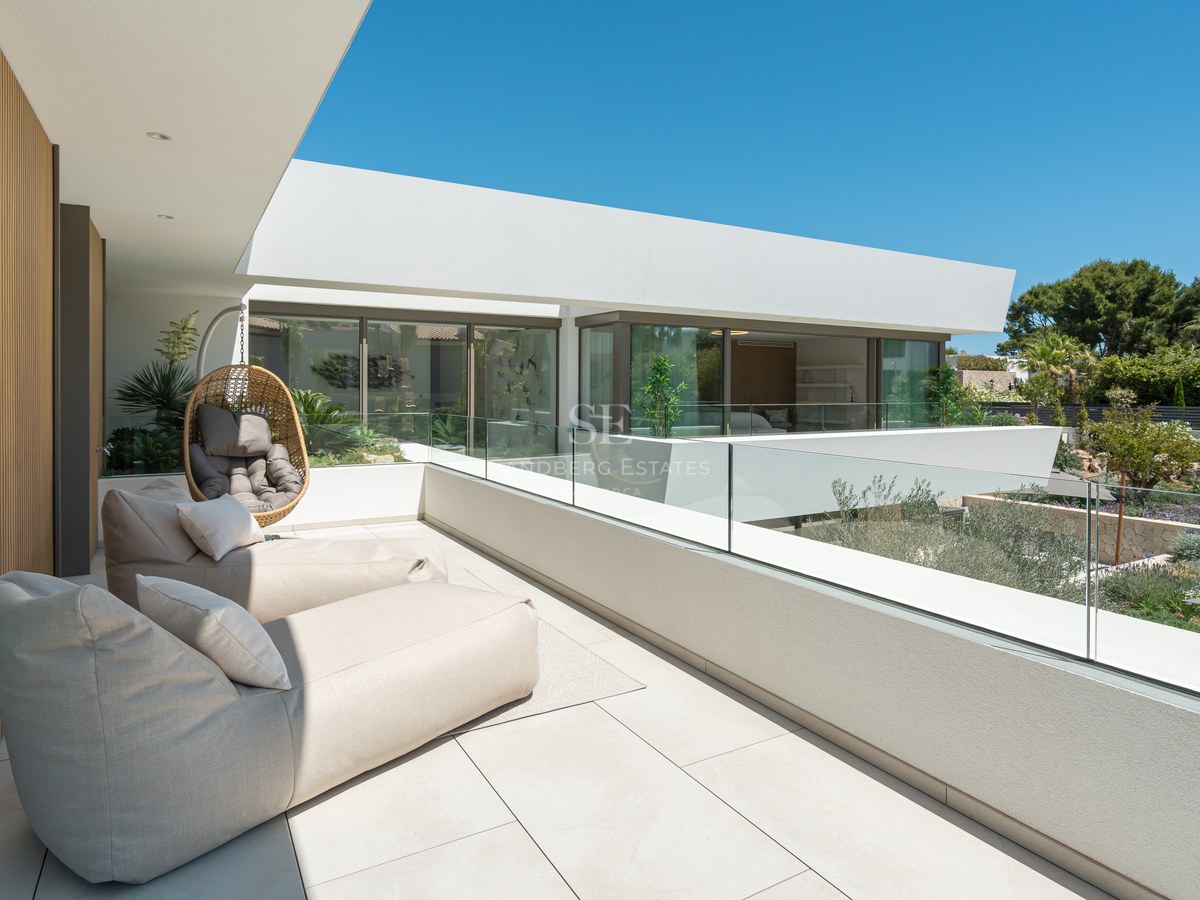 Spacious white terrace with beige lounge chairs, glass railings, and views of a landscaped garden under a clear blue sky.