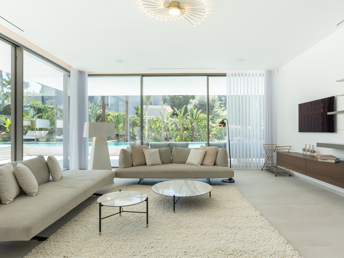 Contemporary living room featuring neutral sofas, marble coffee tables, and large windows overlooking a garden and pool.