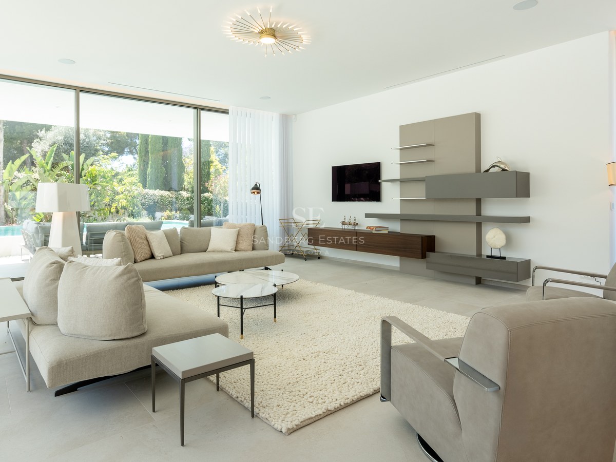 Contemporary living room with beige sofas, marble coffee tables, and floor-to-ceiling glass doors opening to a garden.