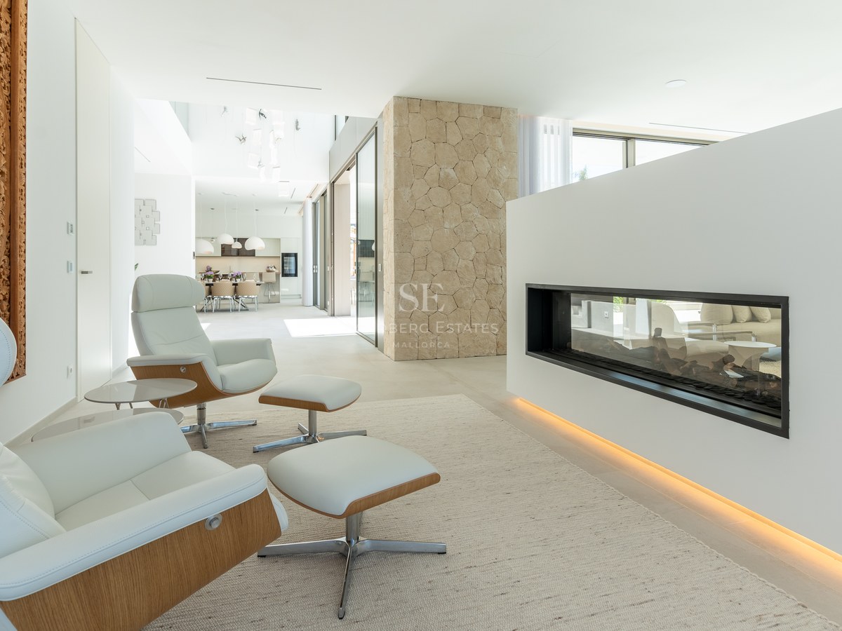 Two white lounge chairs facing a sleek glass fireplace in a bright, modern open-concept living space with stone pillars.