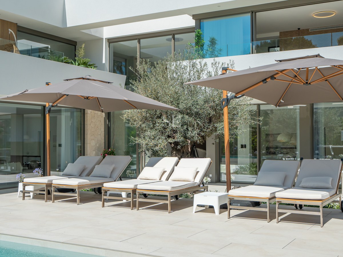 Luxury sun loungers and large umbrellas on a stone terrace beside a pool at a modern white villa.