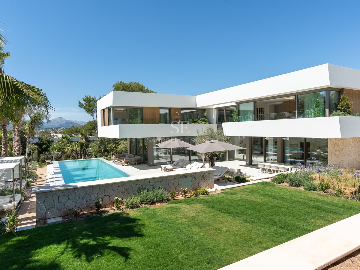 A modern white two-story villa with a turquoise swimming pool, manicured lawn, and Mediterranean garden under a blue sky.