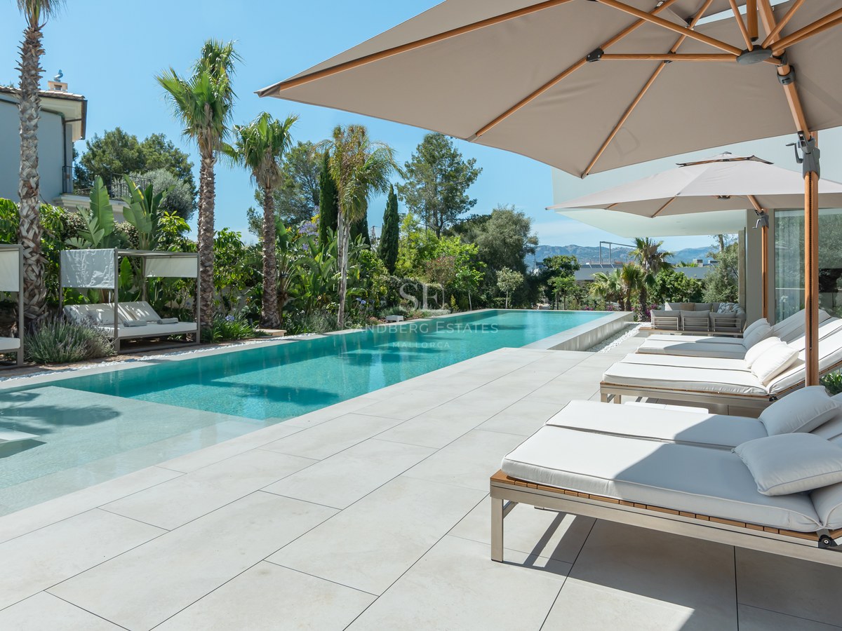 Luxury pool area with white sun loungers, beige umbrellas, and tropical palms under a clear blue sky.