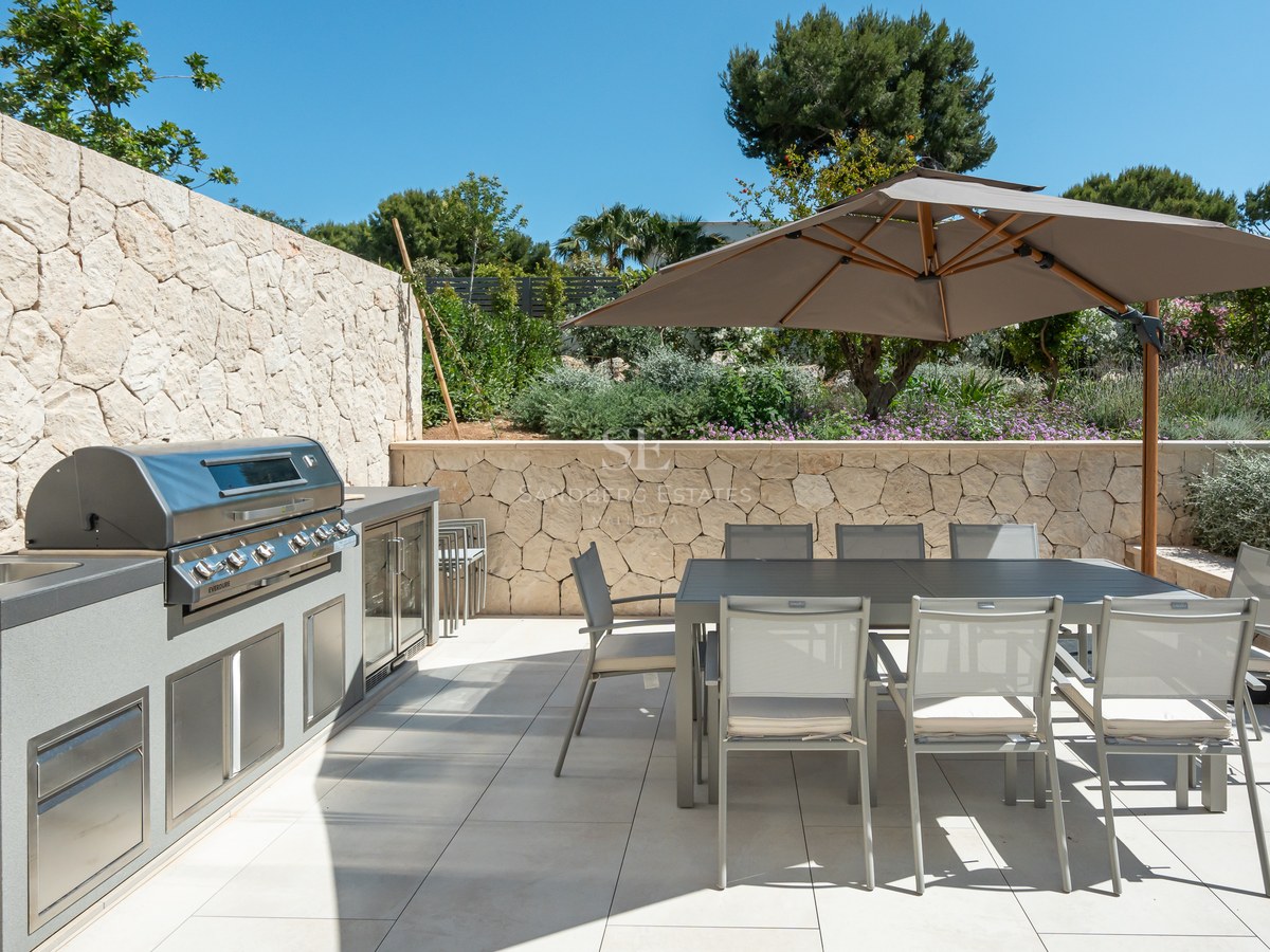 High-end outdoor kitchen with integrated BBQ and 8-seat dining table under a large umbrella on a stone-walled terrace.