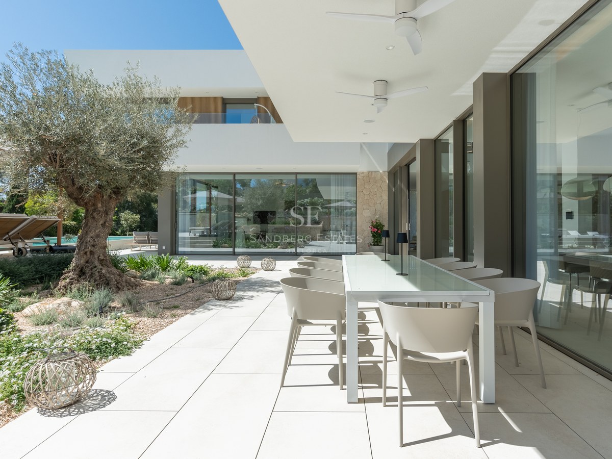 Contemporary villa terrace with white dining table, large glass windows, and a mature olive tree in the garden.
