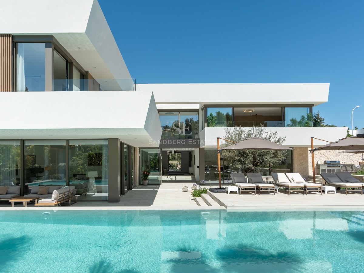 White modern villa with large swimming pool, sun loungers under umbrellas, and floor-to-ceiling glass windows.