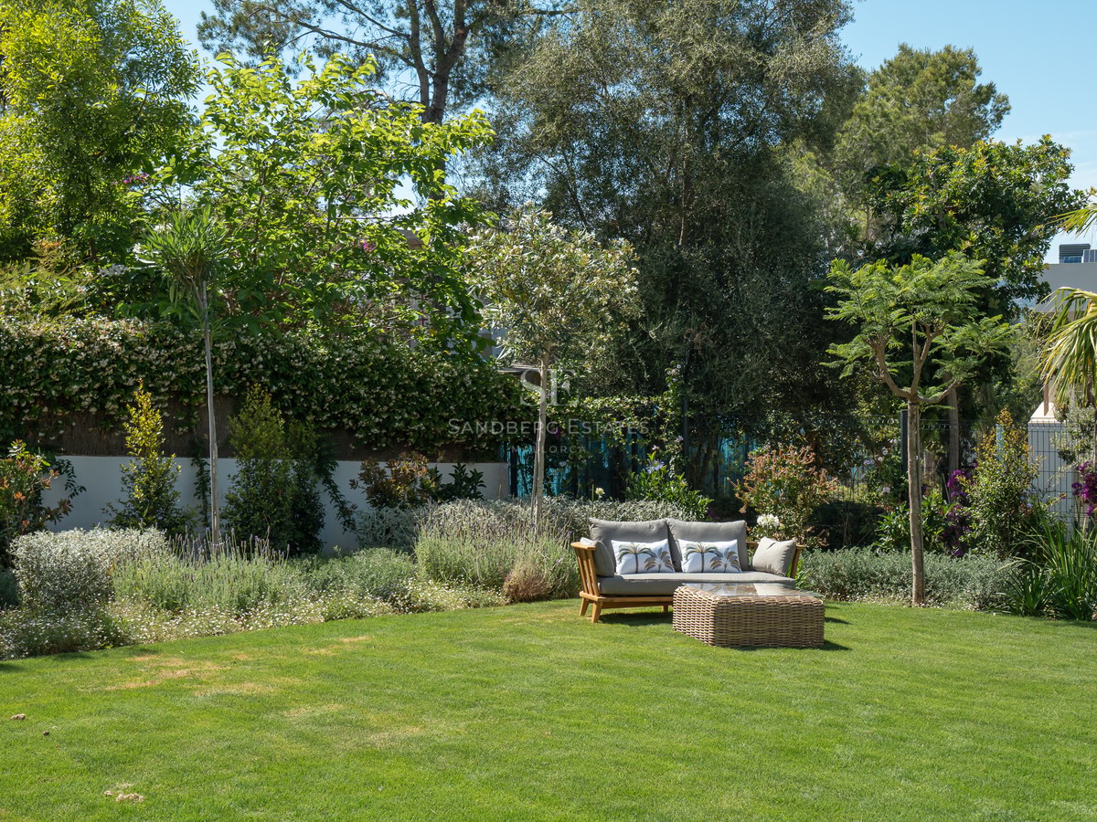 A lush green lawn surrounded by mature Mediterranean trees and flowering shrubs with a wooden sofa and wicker table.