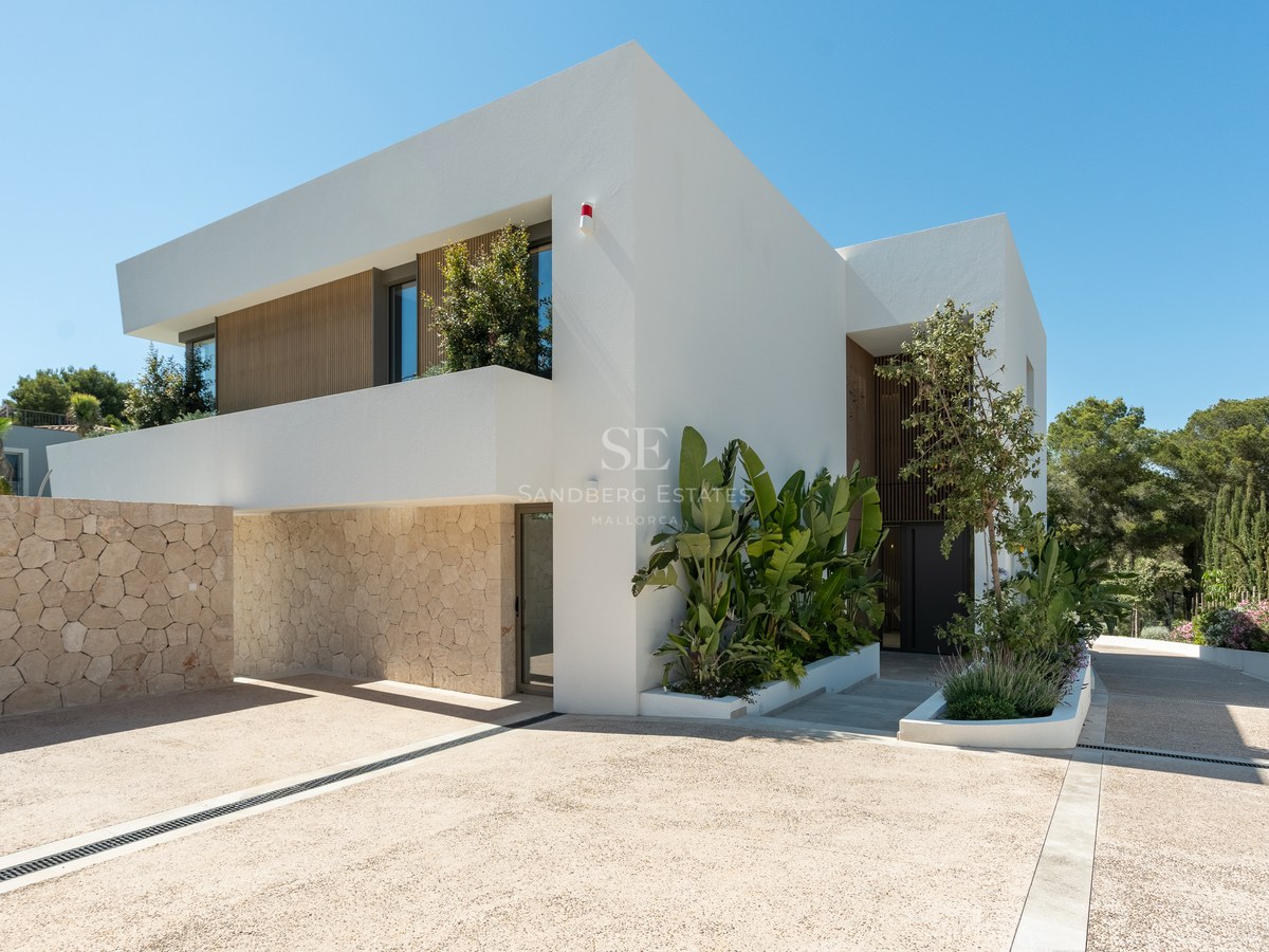 Contemporary two-story white villa with natural stone walls, tropical landscaping, and a spacious paved driveway under a blue sky.