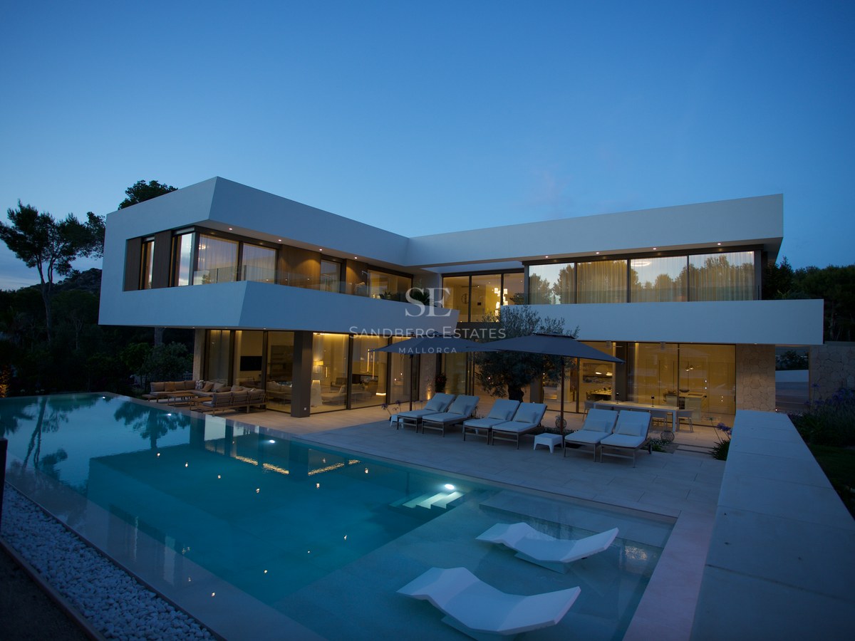 Contemporary two-story white villa with an illuminated infinity pool and submerged loungers under a twilight sky.