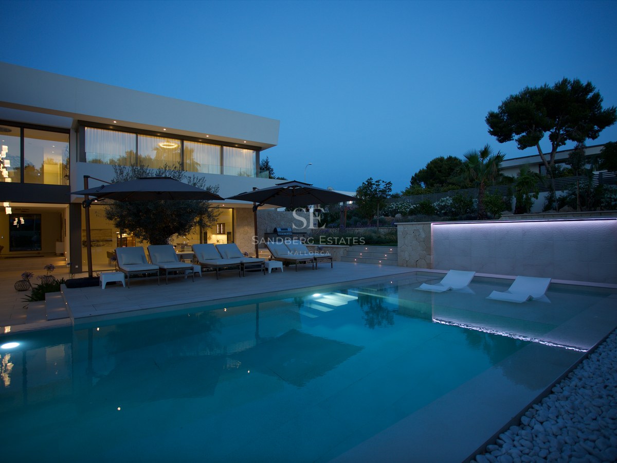 Modern villa exterior at dusk featuring a large swimming pool with integrated loungers and ambient lighting.