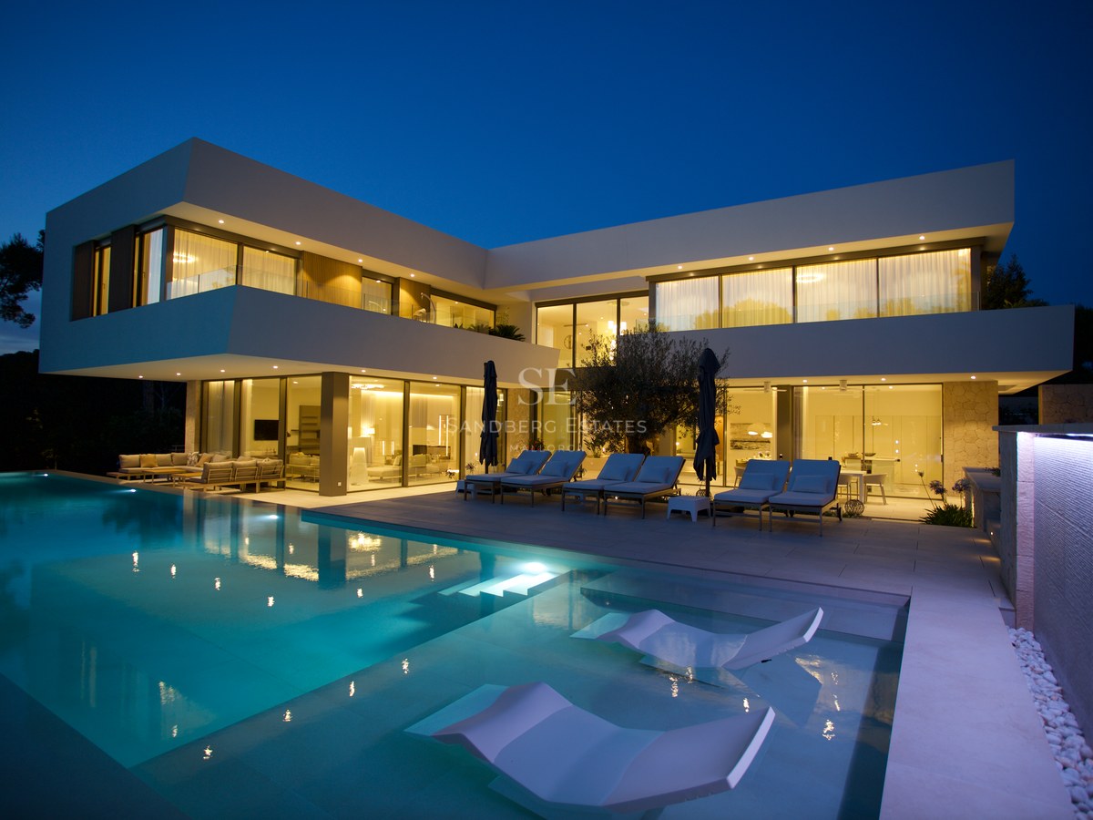 Contemporary white two-story villa at night with a large lit swimming pool, submerged loungers, and minimalist facade.
