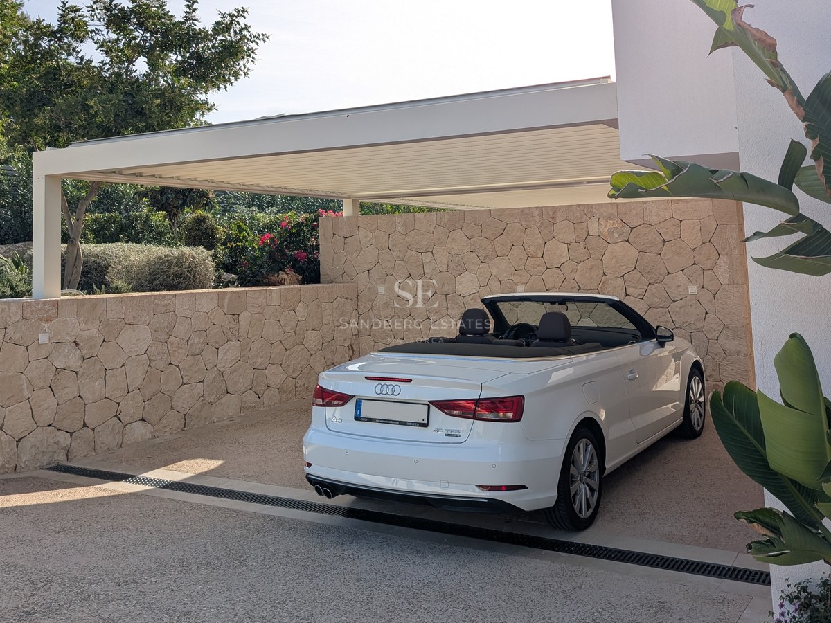 A white convertible parked under a modern louvered pergola, surrounded by natural stone walls and lush greenery.