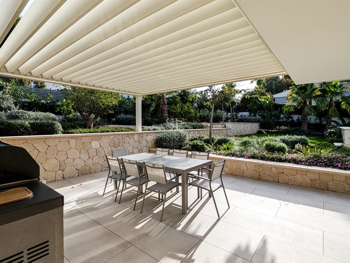 Modern terrace with a large dining table under a white pergola, featuring an outdoor kitchen and stone walls.
