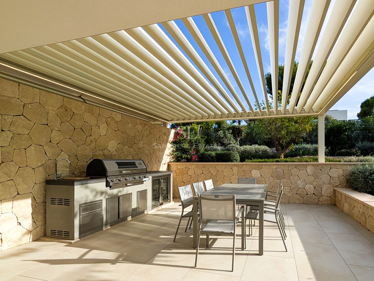 Luxury outdoor kitchen with stainless steel grill and dining table under a louvered pergola with stone walls.