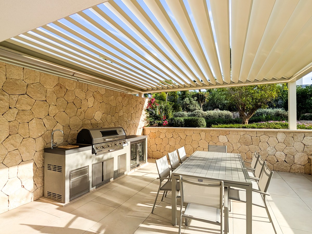 Outdoor kitchen with stainless steel grill and sink under white louvered pergola next to textured stone wall.