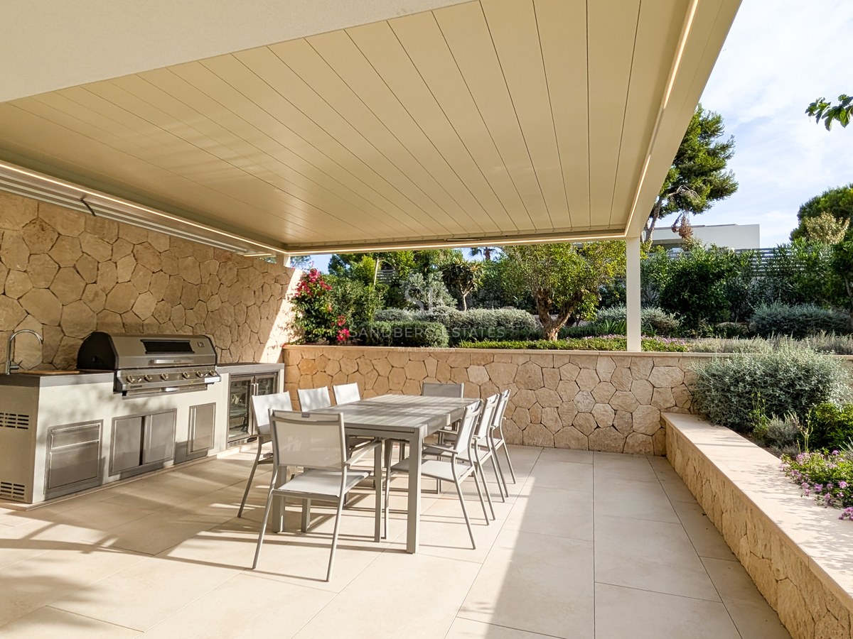 Modern outdoor kitchen with stainless steel BBQ and dining table under a pergola, surrounded by natural stone walls.