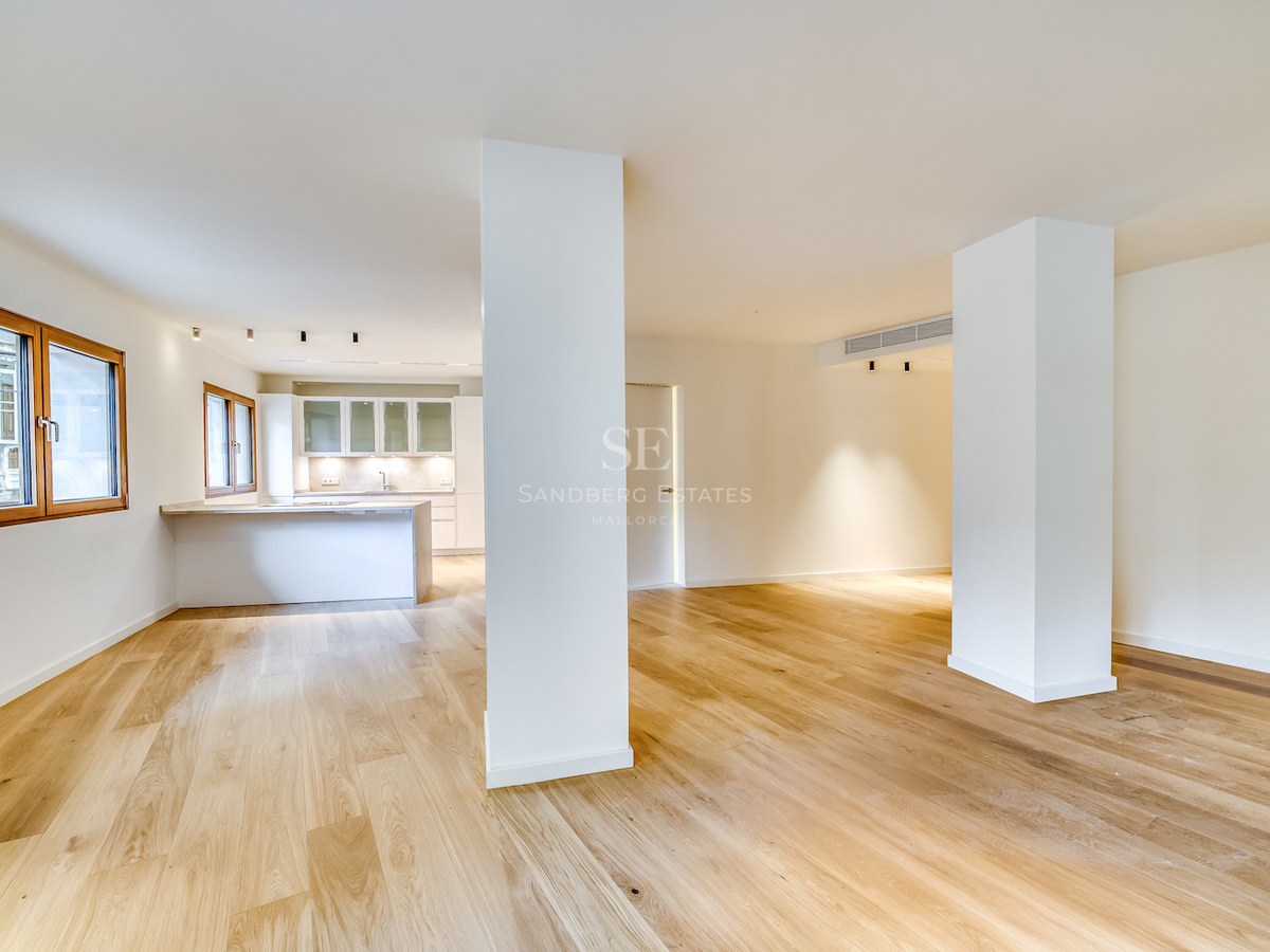 Grand espace de vie ouvert avec parquet en chêne clair, piliers blancs et cuisine minimaliste en arrière-plan.