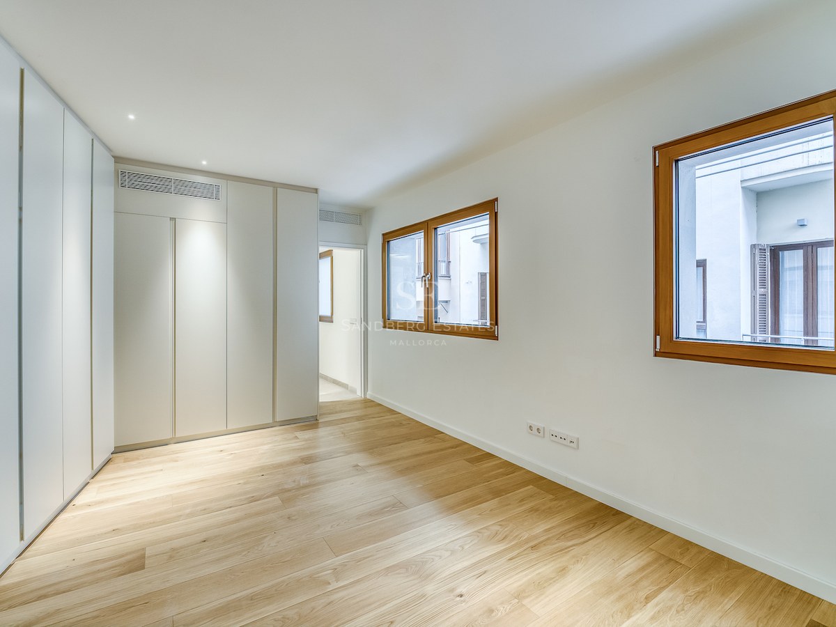Chambre moderne vide avec parquet en chêne clair, placards blancs intégrés et deux fenêtres à cadre en bois.