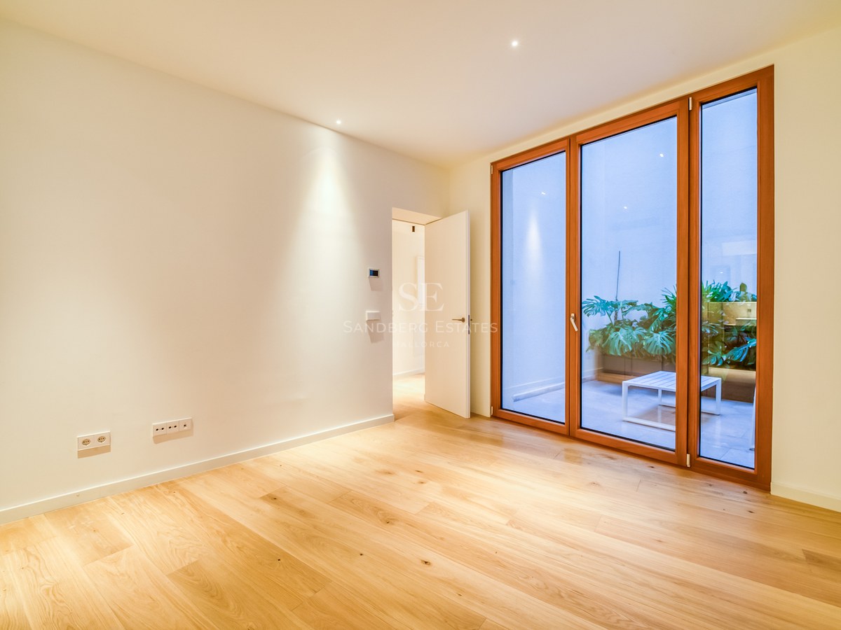 Pièce vide avec parquet en chêne, murs blancs et grandes baies vitrées en bois ouvrant sur un patio arboré.