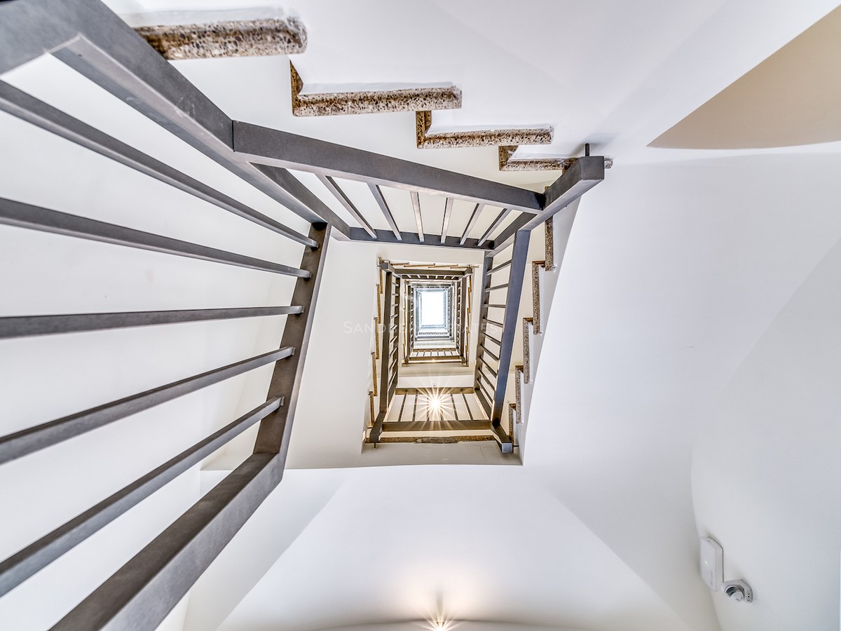 Vue en contre-plongée d'un escalier carré moderne avec garde-corps en métal et murs blancs vers un puits de lumière.