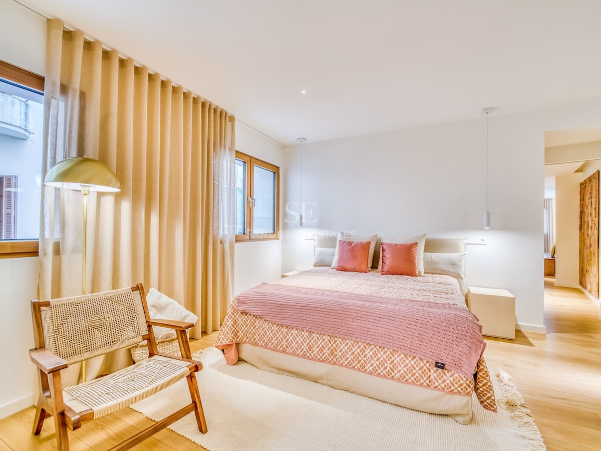 Chambre lumineuse avec lit double, textiles roses, parquet en bois, fauteuil en osier et fenêtres avec rideaux.