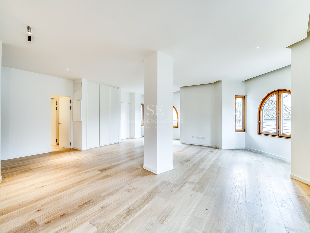 Grande pièce vide avec parquet en chêne clair, murs blancs, pilier central et fenêtres cintrées en bois.