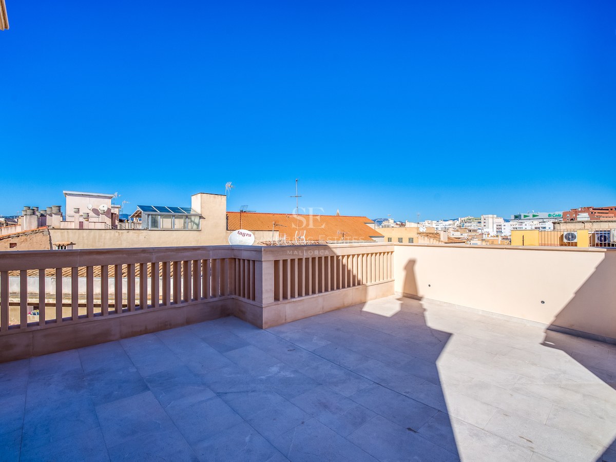 Spacious rooftop terrace with beige stone tiling and a classic balustrade overlooking a Mediterranean city under a blue sky.
