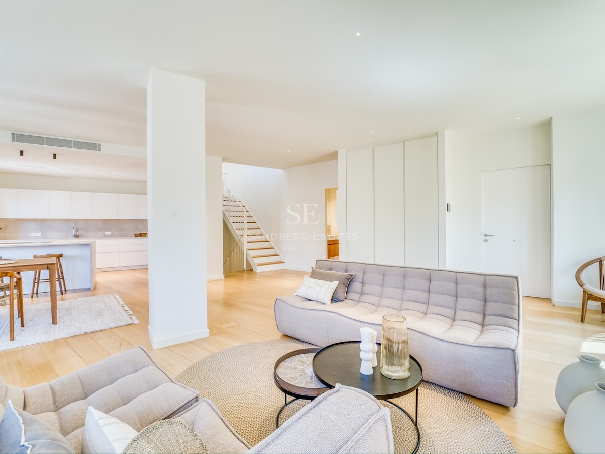 Modern open-plan living room featuring tufted grey sofas, light oak flooring, and a kitchen in the background.