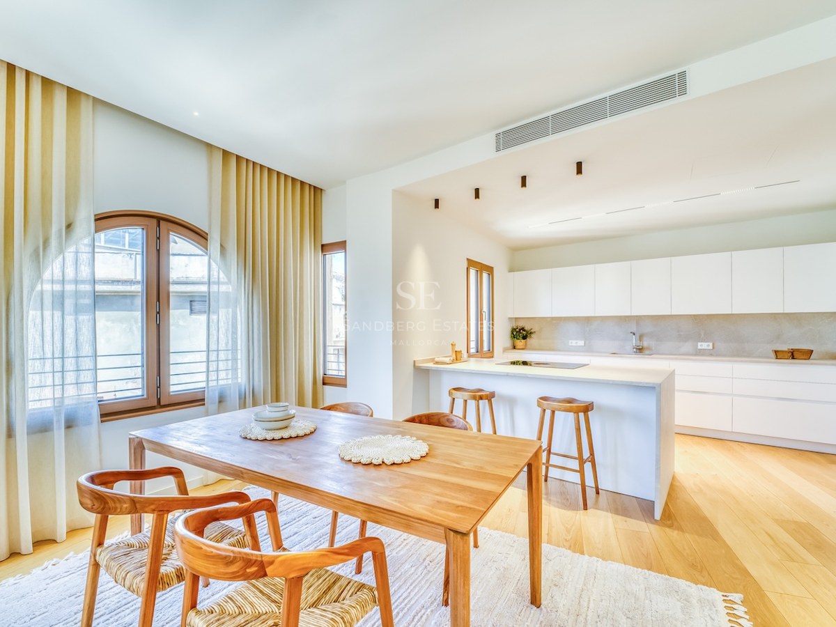 Bright open-plan kitchen with white cabinetry and a wooden dining table under an arched window with light curtains.