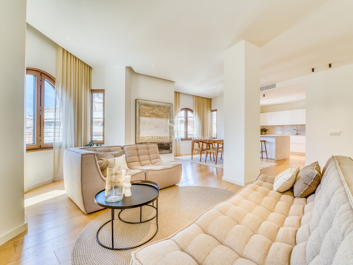 Bright living room with beige sofas, oak flooring, and arched windows leading to a dining area and kitchen.