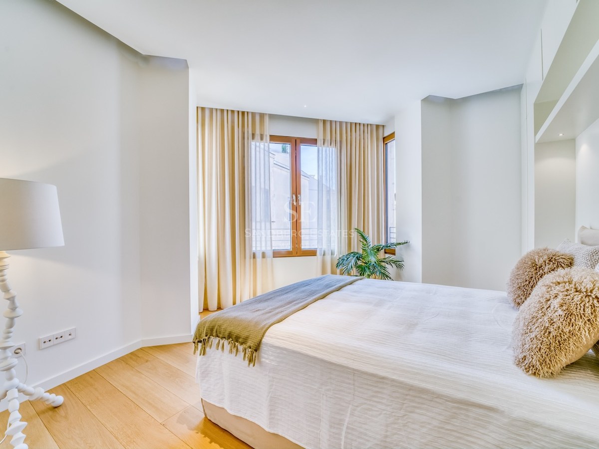 A bright bedroom with light oak floors, a double bed with textured pillows, and a large window with sheer curtains.
