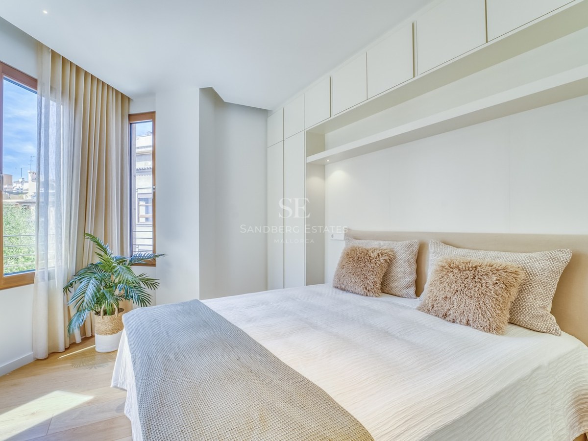 Bright master bedroom featuring integrated white storage, wooden window frames, and neutral textiles.